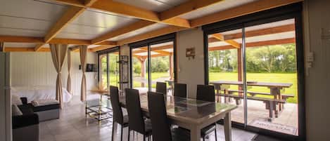 A dining area with a glass table and chairs, a sofa, and a view of a garden through large windows.