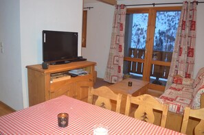 A room with a television on a wooden stand, a table with a checkered tablecloth, and a window with patterned curtains. - Apartment for 6 People in Valmeinier (Valmeinier)