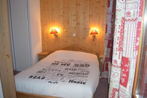 A bedroom with a wooden headboard, two bedside lamps, a bed with a white sheet, and a wall with a decorative pattern. - Apartment for 6 People in Valmeinier (Valmeinier)