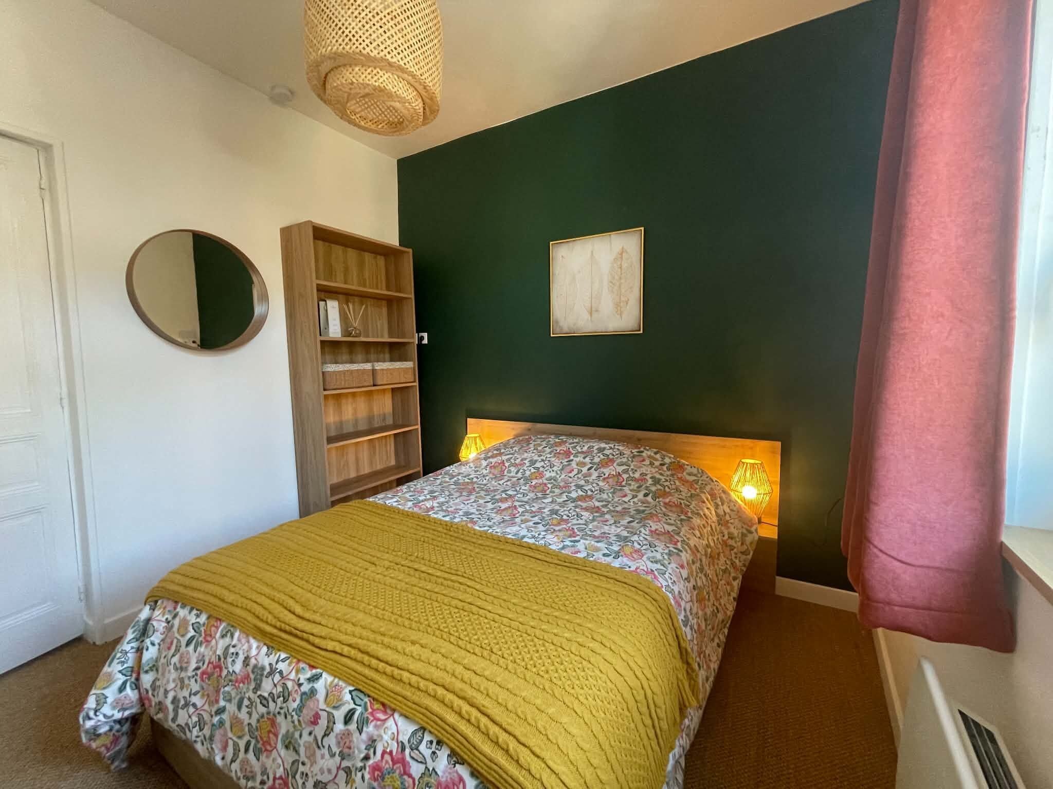 A bedroom with a bed, a wooden bookshelf, a round mirror, a framed picture, and a window with a curtain.