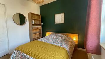 A bedroom with a bed, a wooden bookshelf, a round mirror, a framed picture, and a window with a curtain.