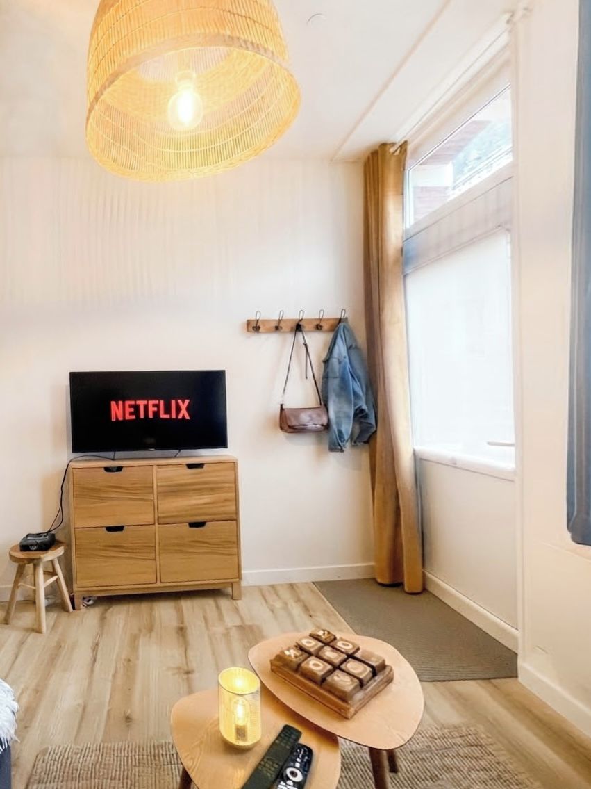 Una habitación con una televisión mostrando 'NETFLIX', un armario de madera, un taburete pequeño, una mesa con un control remoto y un perchero.