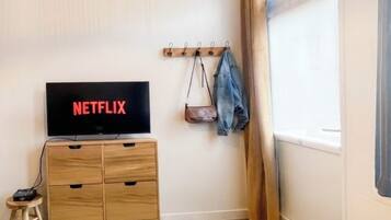 Una habitación con una televisión mostrando 'NETFLIX', un armario de madera, un taburete pequeño, una mesa con un control remoto y un perchero.