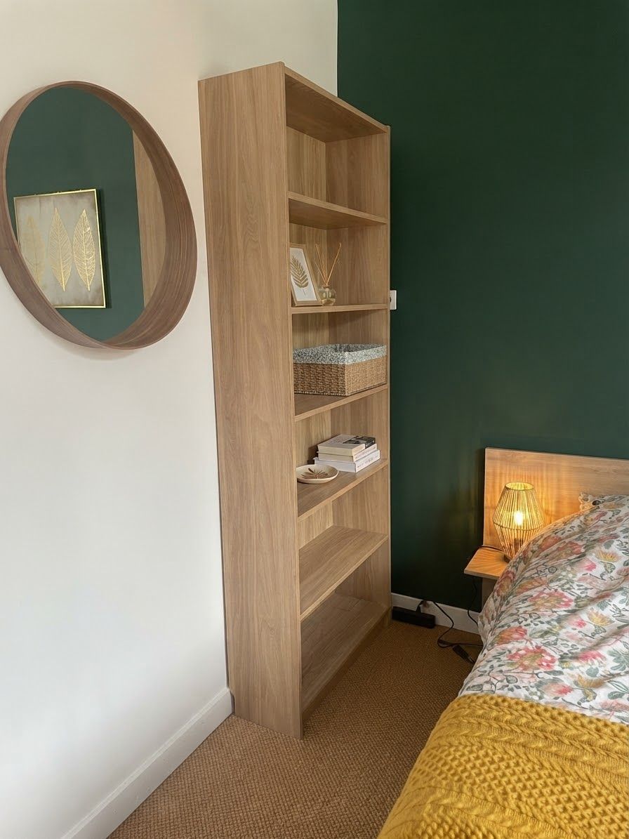 A bedroom with a wooden bookshelf, a round mirror, a bed with a floral bedspread, and a bedside lamp.