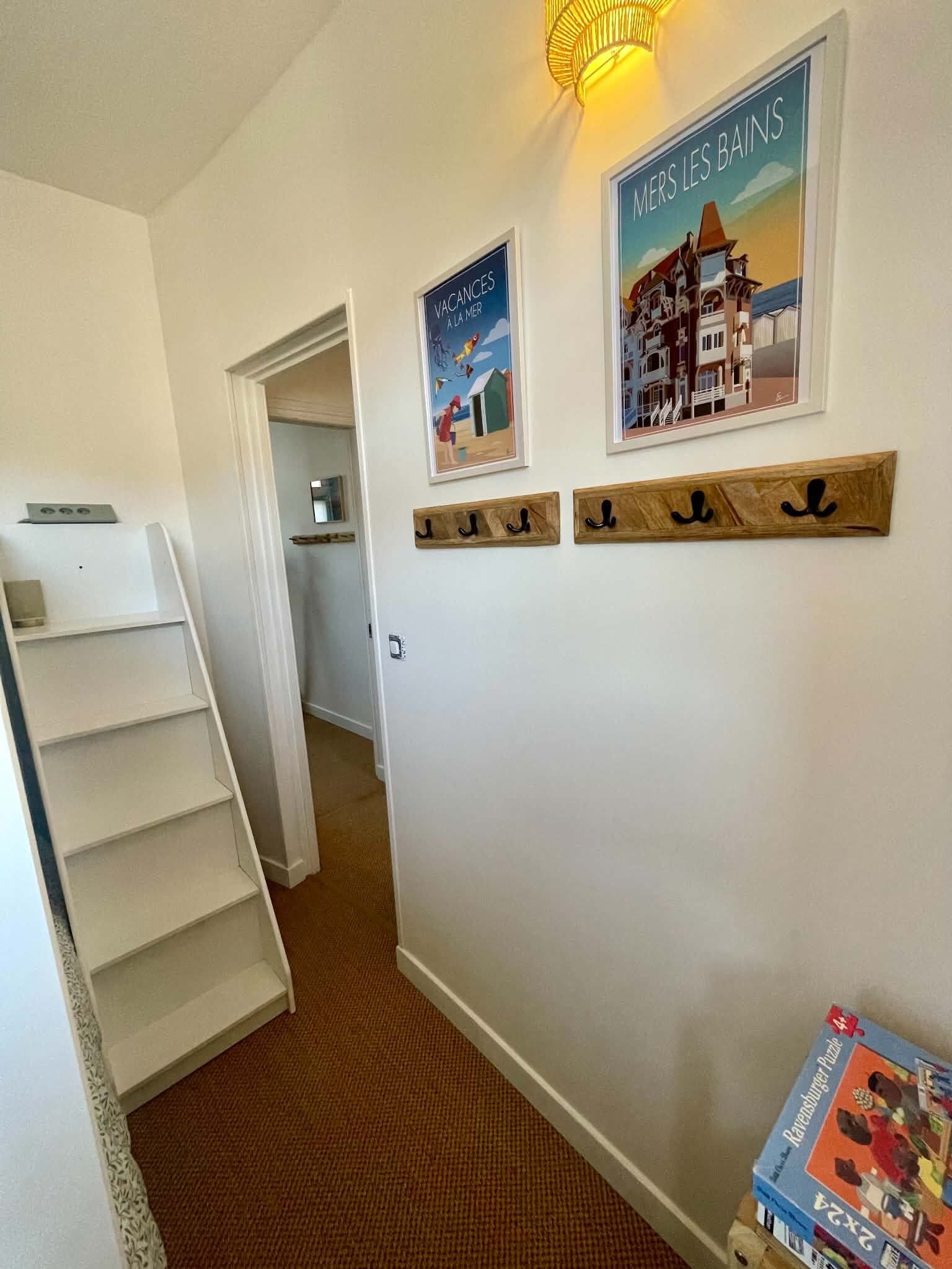 A hallway with a wooden coat rack, a framed poster of a seaside town, and a bookshelf with a puzzle box.