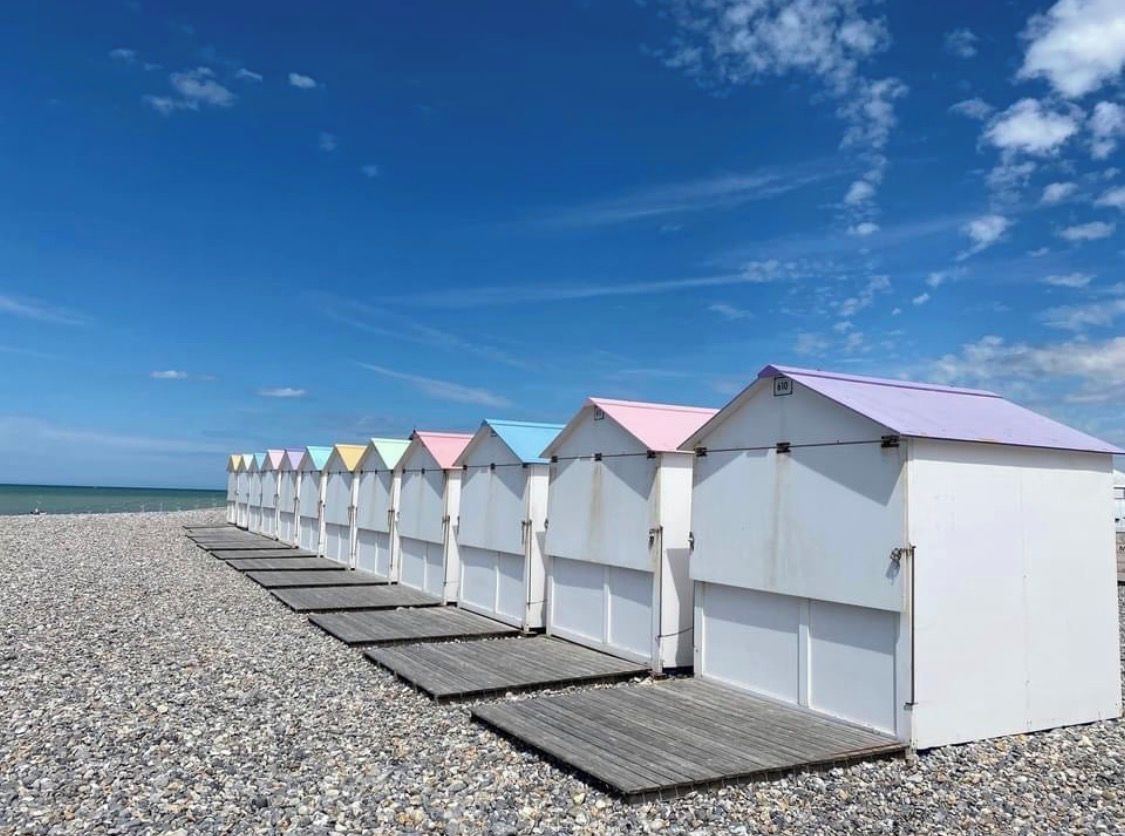 A row of beach huts with a wooden deck in front, set on a pebbled beach with a clear sky.