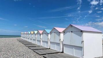 A row of beach huts with a wooden deck in front, set on a pebbled beach with a clear sky.
