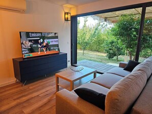 Ein modernes Wohnzimmer mit einem großen Flachbildfernseher, einer Couch, einem Couchtisch und Blick auf einen Garten durch große Fenster.