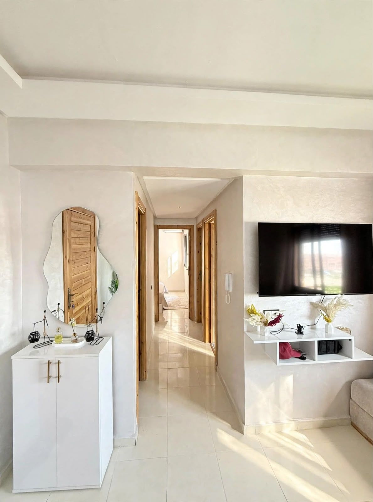 A modern interior with a hallway leading to a bedroom, a white vanity with a mirror, a mounted TV, and a shelf with decorative items.