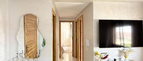 A modern interior with a hallway leading to a bedroom, a white vanity with a mirror, a mounted TV, and a shelf with decorative items.