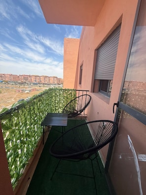 A balcony with a table and chairs, a window with a shutter, and a view of buildings in the distance. - APARTMENT 3 MINUTES FROM THE AIRPORT (marrakech)