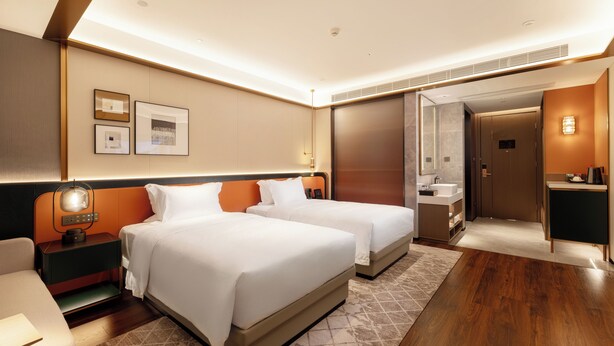 Ein Hotelzimmer mit zwei Betten, einem Schreibtisch, einem Stuhl, einem Nachttisch, einer Lampe, einem Wandspiegel, einer Badezimmertür und einem kleinen Tisch mit einer Tasse.