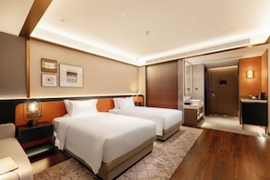 A hotel room with two beds, a desk, a chair, a nightstand, a lamp, a wall-mounted mirror, a door to the bathroom, and a small table with a cup. - FURAMA SHANSHUI HOTEL (Jiangmen)