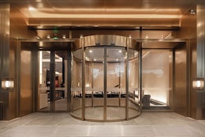 Una puerta giratoria con paneles de vidrio y estructura metálica, que da acceso a un vestíbulo con mostrador de recepción y asientos.