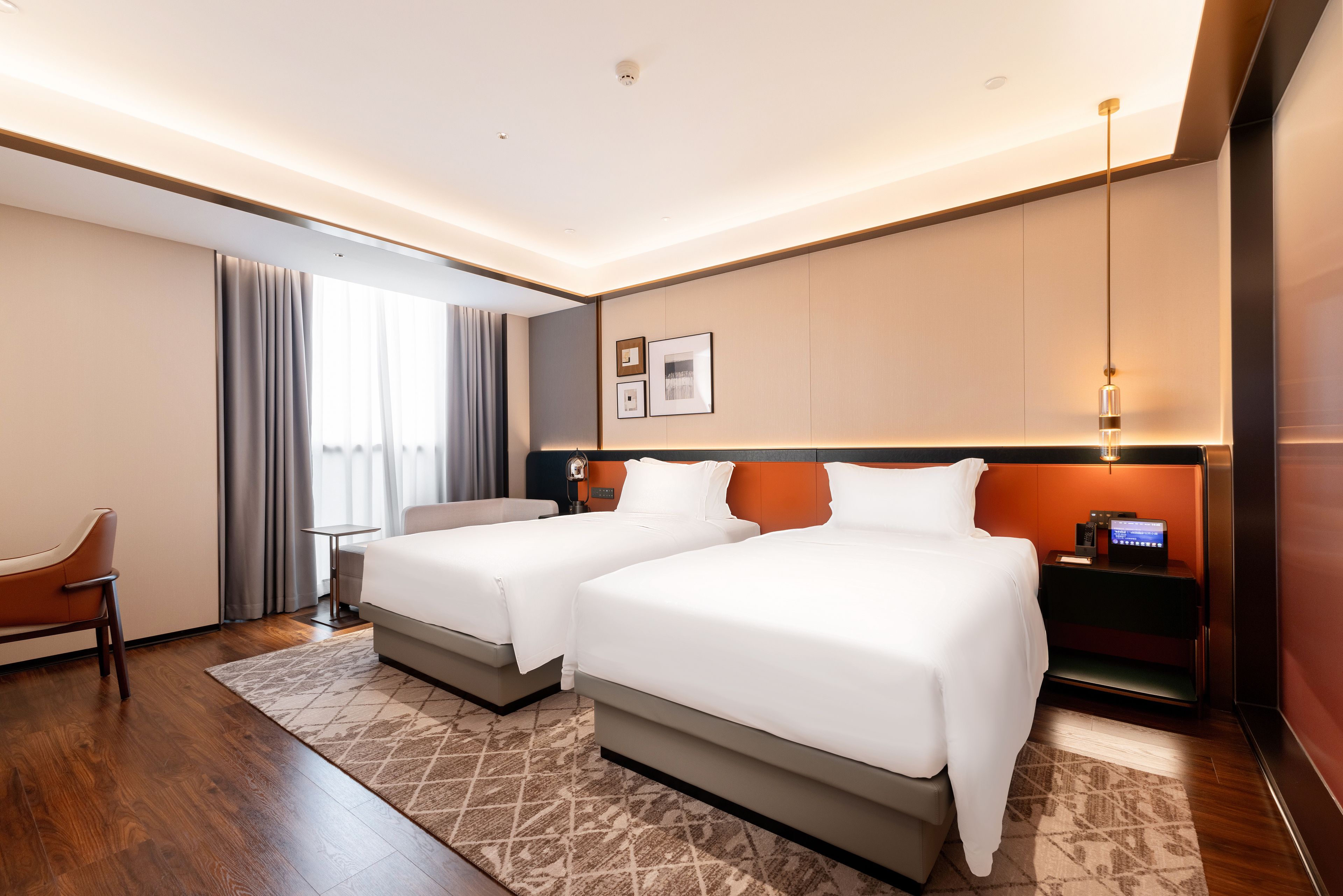 Habitación de hotel con dos camas, un escritorio, una silla, un ventanal grande con cortinas y un televisor instalado en la pared.