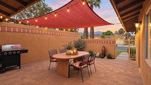 Un patio exterior con una parrilla, una mesa con sillas y luces colgantes.