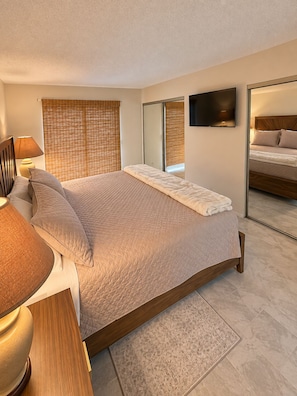 Un dormitorio con cama, mesita de noche, lámpara, espejo y ventana con persianas.