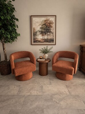 Two armchairs with a round table between them, a framed painting on the wall, and a potted plant to the left.