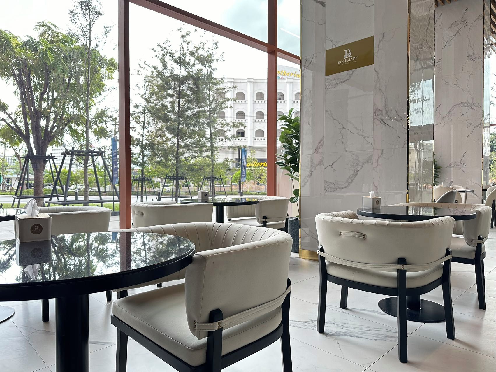A modern indoor seating area with round tables and chairs, large windows offering a view of the street and trees, and a marble wall with a sign.
