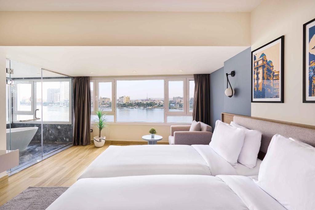 Habitación de hotel con una cama grande, un sofá, una mesita y vistas a un paisaje urbano a través de amplios ventanales.