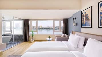 Habitación de hotel con una cama grande, un sofá, una mesita y vistas a un paisaje urbano a través de amplios ventanales.