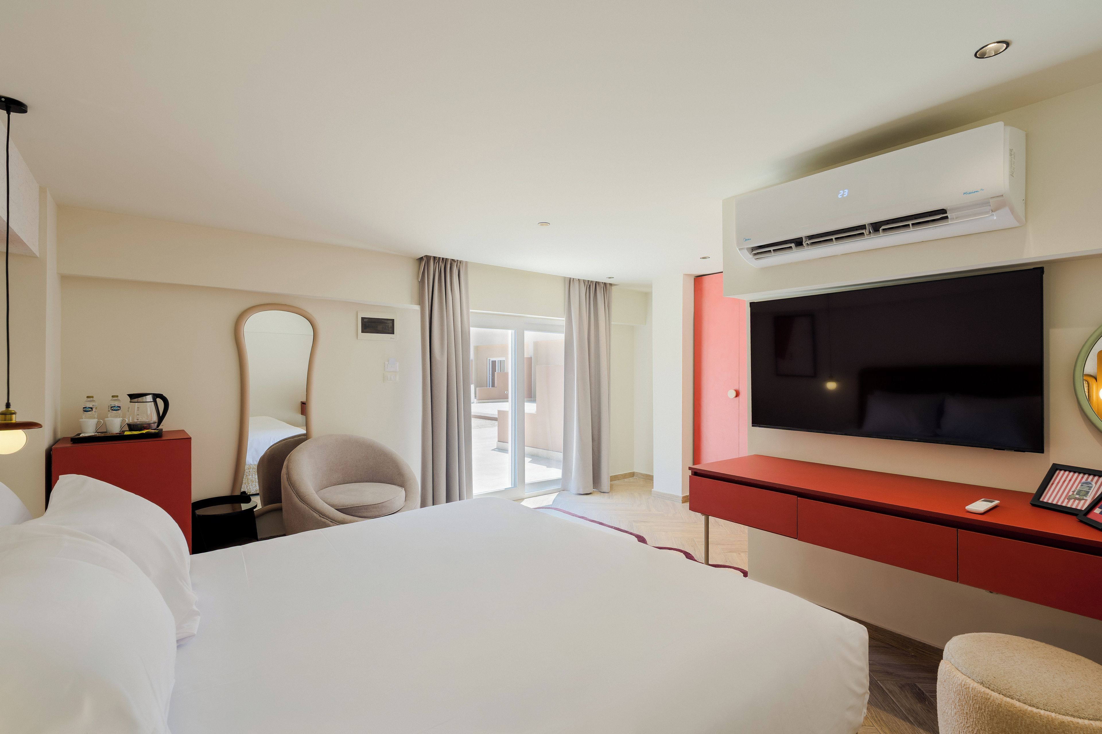 A hotel room with a bed, a TV, an air conditioning unit, a desk, a chair, and a mirror.