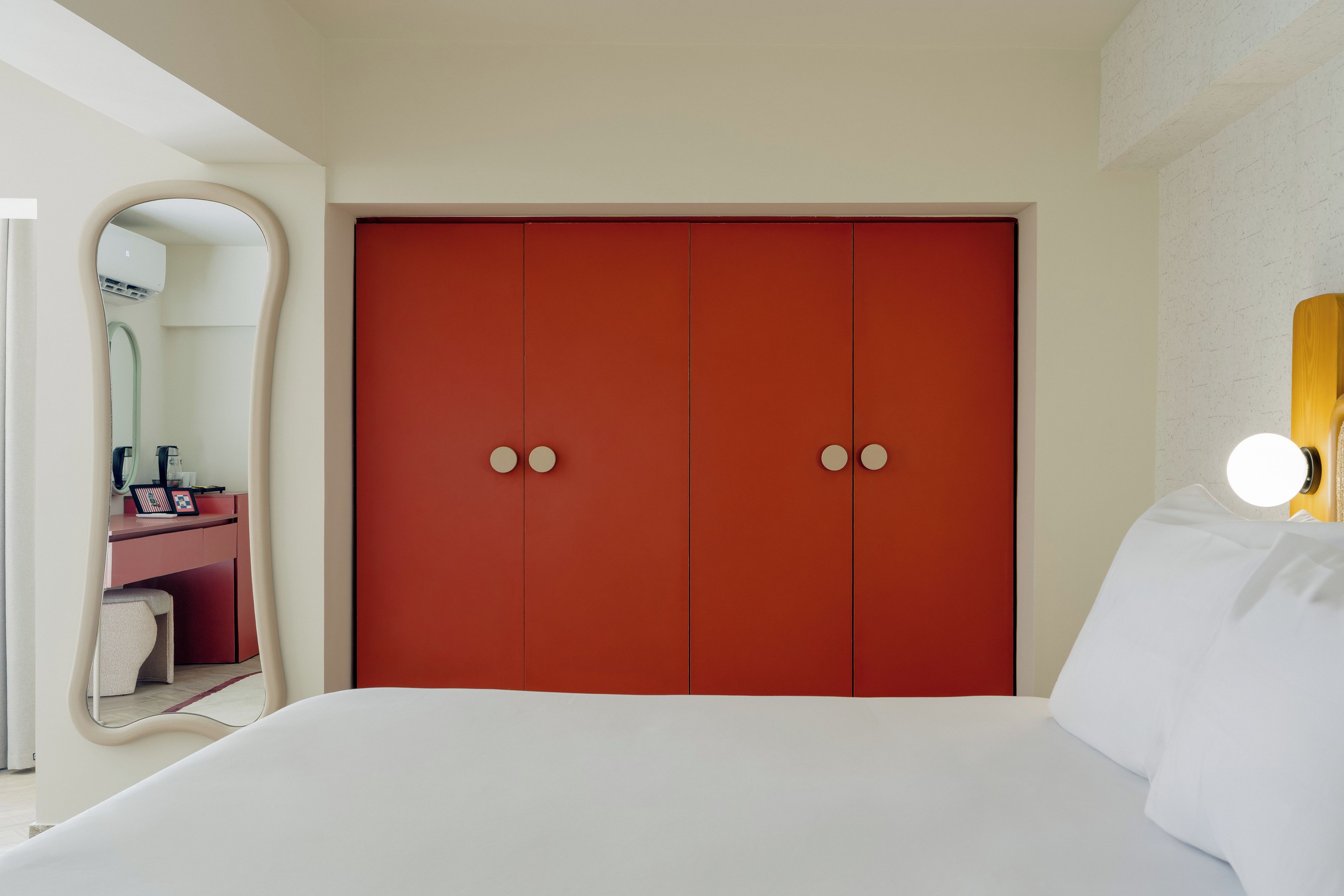 A hotel room with a large bed, a red wardrobe, a desk with a chair, and a mirror.