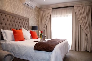A hotel room with a tufted headboard, a bed with white and orange bedding, a bedside lamp, and a window with curtains. - Classique  Grace Boutique Hotel and Spa (Morningside Manor)