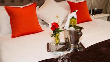 Un letto con lenzuola bianche, due cuscini rossi, un comodino con una bottiglia, un bicchiere e un vaso con una rosa rossa.