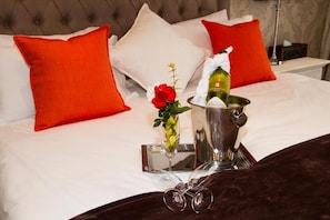 A bed with white linens, two red pillows, a bedside table with a bottle, a glass, and a vase with a red rose. - Classique  Grace Boutique Hotel and Spa (Morningside Manor)
