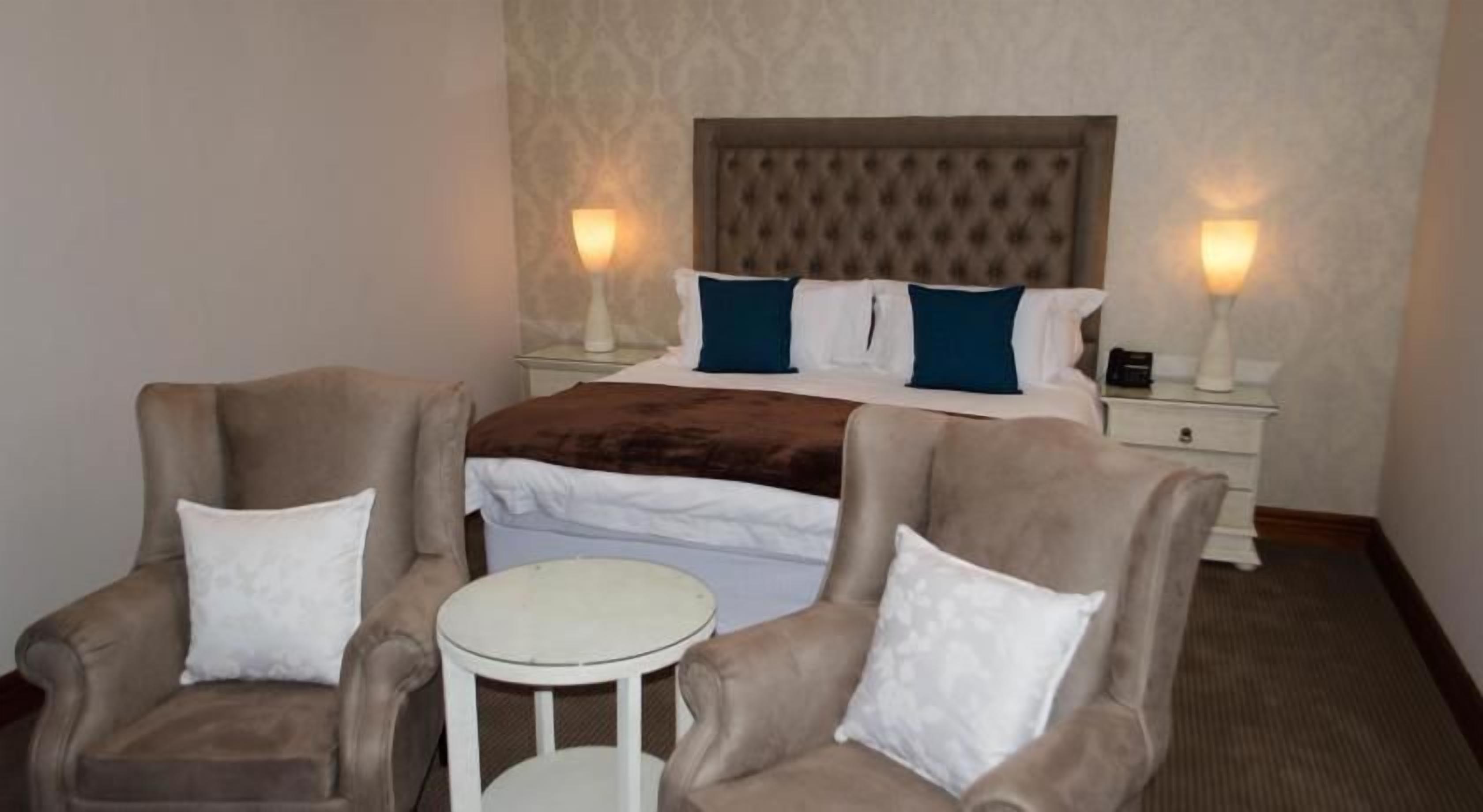 Una habitación de hotel con una cama, dos sillones, una mesita redonda y dos lámparas de noche.