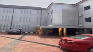 A building with a covered entrance, a car parked in front, and a person standing near the entrance. - Asabana Suites and Hotels Enugu (Enugu)