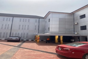 Un edificio con un vestíbulo cubierto, un coche estacionado adelante y una persona parada cerca de la entrada.
