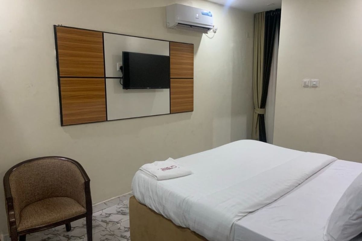 Habitación de hotel con cama, una silla, un televisor de pantalla plana montado en un panel de madera y un equipo de aire acondicionado.