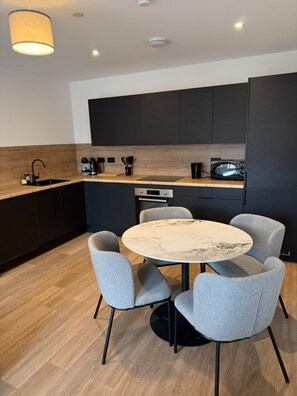 A modern kitchen with dark cabinetry, a round marble table, and grey upholstered chairs. - Standard Two Bedroom Apartment Near MediaCity (Salford)