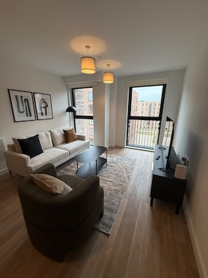 A modern living room with a sofa, armchair, coffee table, and a TV stand. There are large windows providing views of the outside. - Standard Two Bedroom Apartment Near MediaCity (Salford)