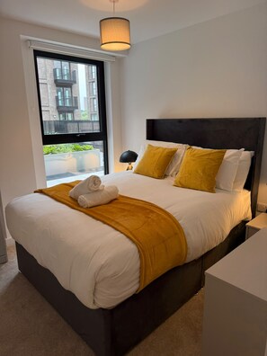 Una habitación de hotel moderna con una cama grande, una ventana con vistas a edificios, una lámpara de noche y una cama bien hecha con una toalla.