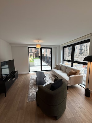A modern living room with a sofa, armchair, and TV, large windows, and a view of buildings.