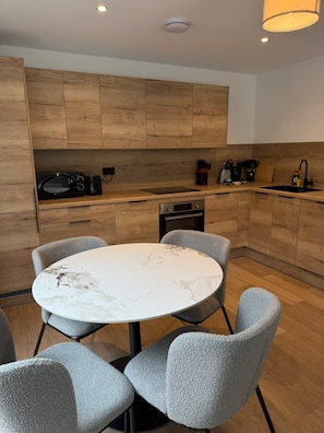 A modern kitchen with wooden cabinets, a round marble table, and grey upholstered chairs.