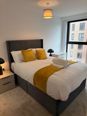 Een net opgemaakt bed met wit en geel bedlinnen, een donker hoofdbord en een bedlampje.