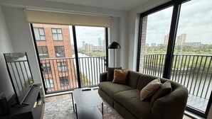 Ein modernes Wohnzimmer mit Sofa, Sessel und Couchtisch. Ein großes Fenster bietet Blick auf Gebäude und einen Balkon.