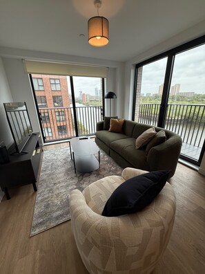 Una sala de estar moderna con un sofá, un sillón y una mesa de centro. Hay un ventanal con vistas a edificios y un balcón.