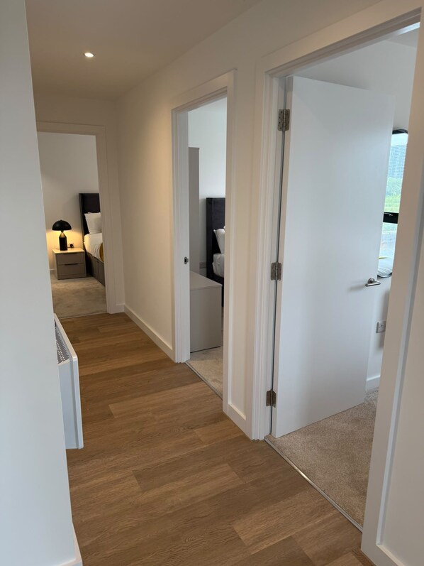 A hallway with wooden flooring, white walls, and a door leading to a bedroom with a bed and nightstand.