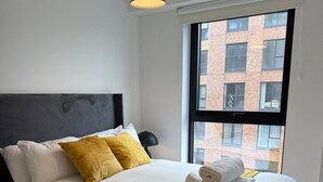 Ein ordentlich bezogenes Bett mit weißen Leinentüchern und einer senffarbenen Tagesdecke, ein schwarzes Kopfteil und ein Fenster mit Blick auf ein Backsteingebäude.