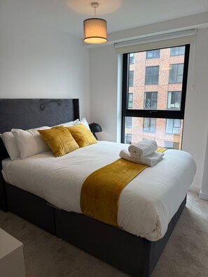 Una cama bien hecha con sábanas blancas y un plaid color mostaza, cabecera negra y una ventana con vistas a un edificio de ladrillo.