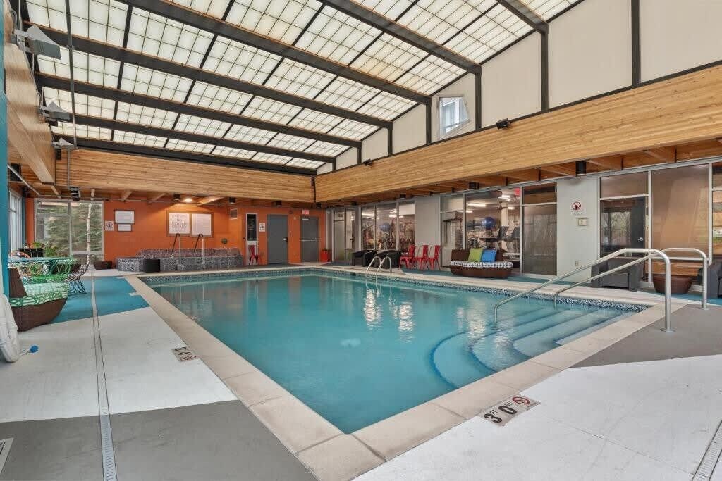 An indoor swimming pool with a glass ceiling, seating area, and a view of the city.