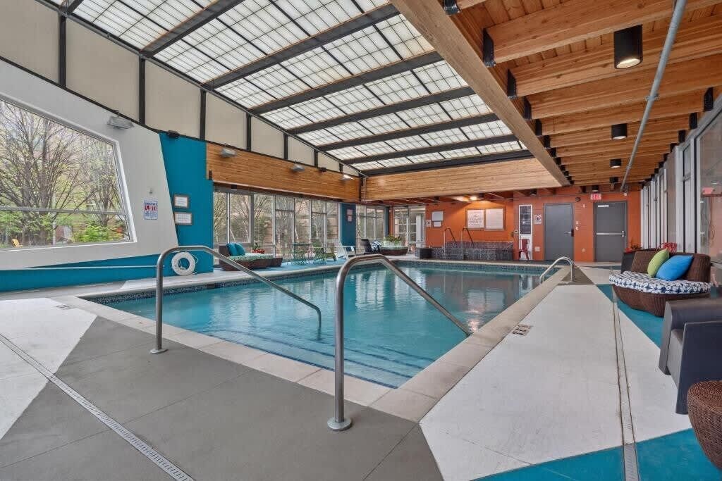 An indoor swimming pool with a glass ceiling, wooden beams, and a lounge area with seating.