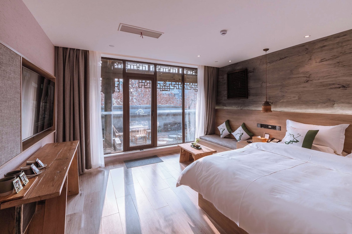 Quarto de hotel com uma cama grande, uma escrivaninha de madeira, vista através de uma porta de vidro e uma luminária moderna.
