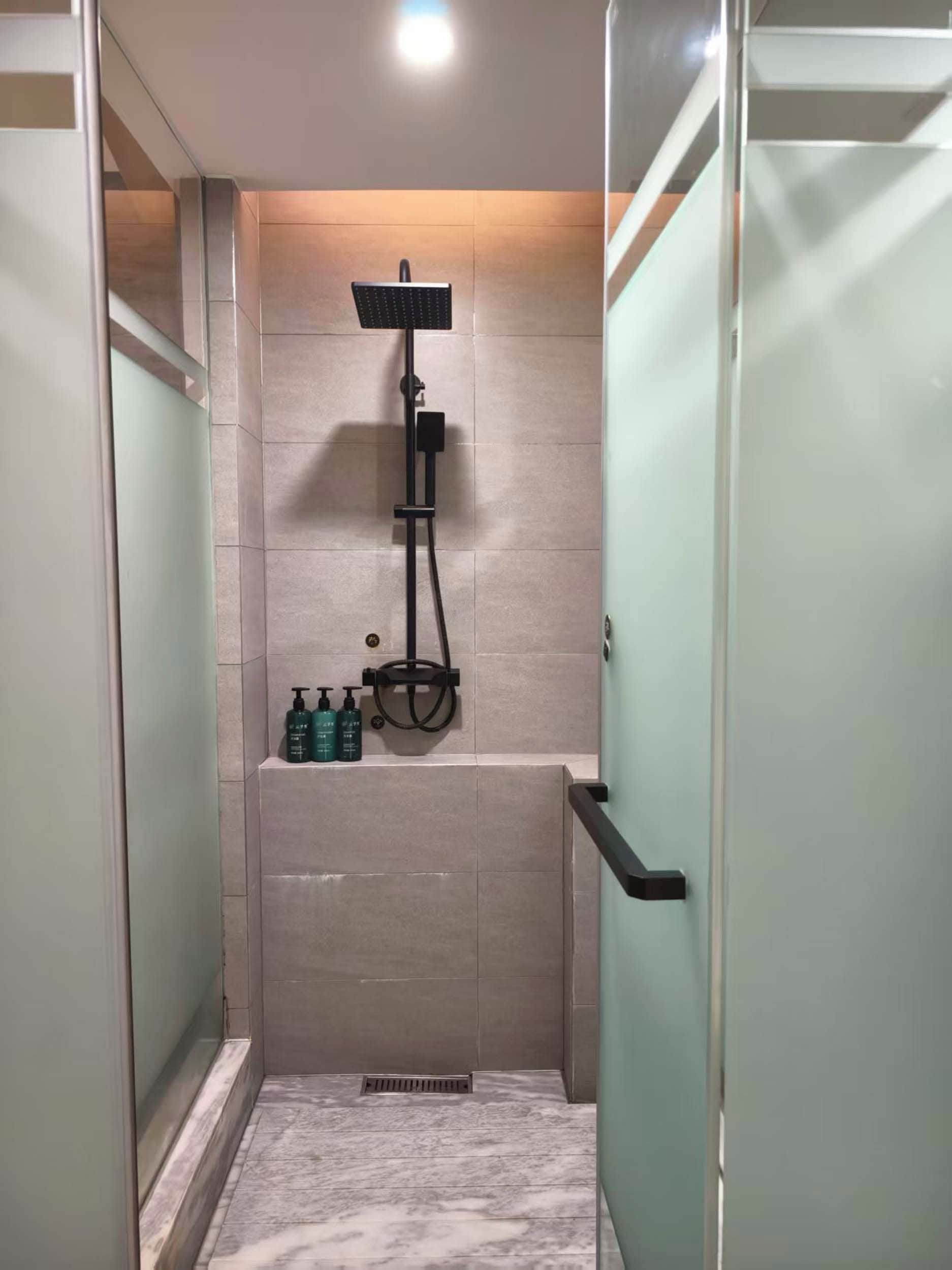 A glass-enclosed shower stall with a handheld showerhead, a handheld shower attachment, and a bottle of shampoo on the wall.