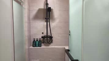 A glass-enclosed shower stall with a handheld showerhead, a handheld shower attachment, and a bottle of shampoo on the wall.